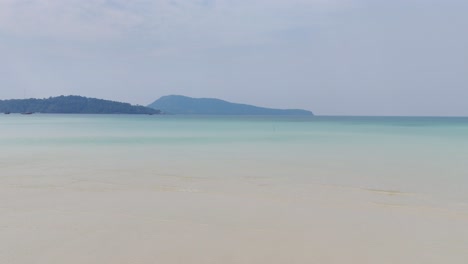 serene seascape on kaoh rong sanloem, cambodia, with soft blue-green gradient waters meeting pale sandy shallows and distant lush hills rising on the horizon under a hazy sky, drone low shot