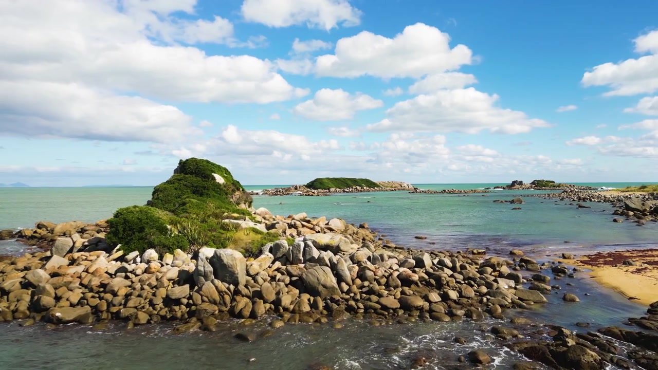 Luftfoto over stenstrand med klipper og små øer i havet under blå himmel.
