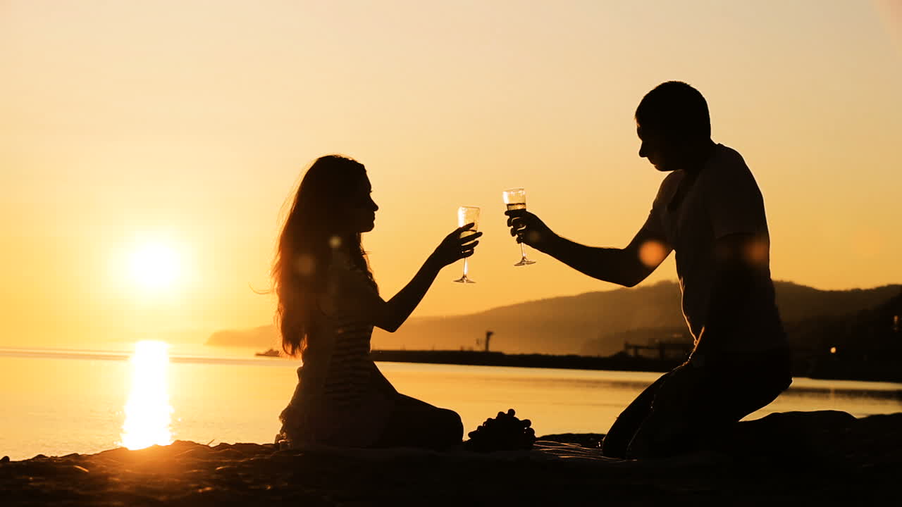 Romantic relationships concept. Silhouettes of couple sitting and drinking wine on beach at sunset