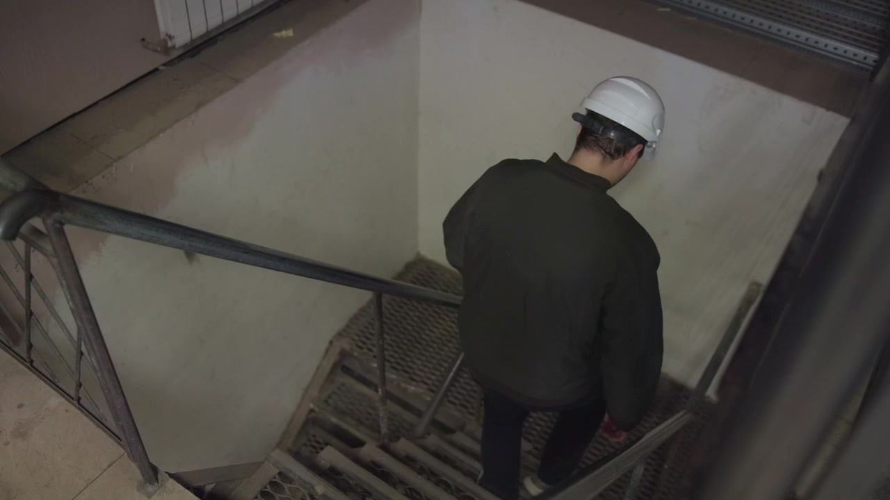Operaio con cappello bianco e giacca scura scende scale metalliche a griglia all'interno di uno stretto pozzo di servizio, afferrando il corrimano accanto a una parete bianco sporco usurata, con luce attenuata, texture ruvide, confinato.