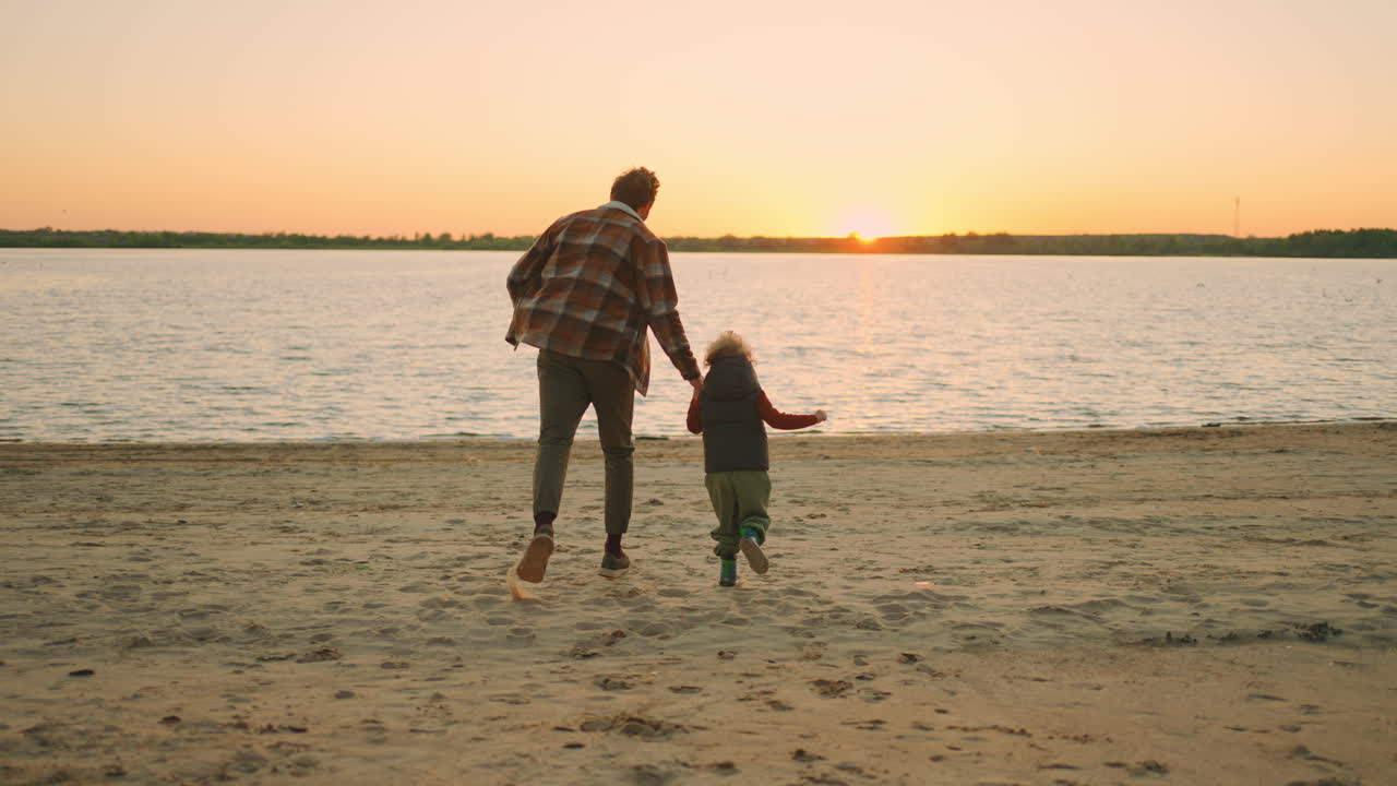Far og søn holder i hånd og løber mod floden ved solnedgang. Lykkelig familieweekend.