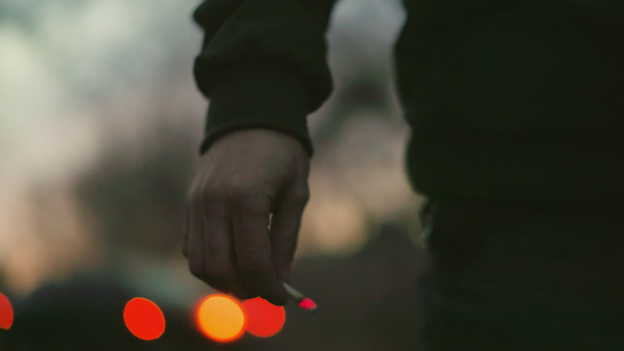 불타는 담배를 들고 있는 남자의 손의 근접한 모습, 반이는 끝이 흐릿한 배경에 생생한 은 점을 만니다.