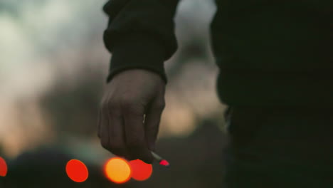 a close-up view of a man's hand holding a lit cigarette, the glowing tip creating a vivid red point against the blurred background
