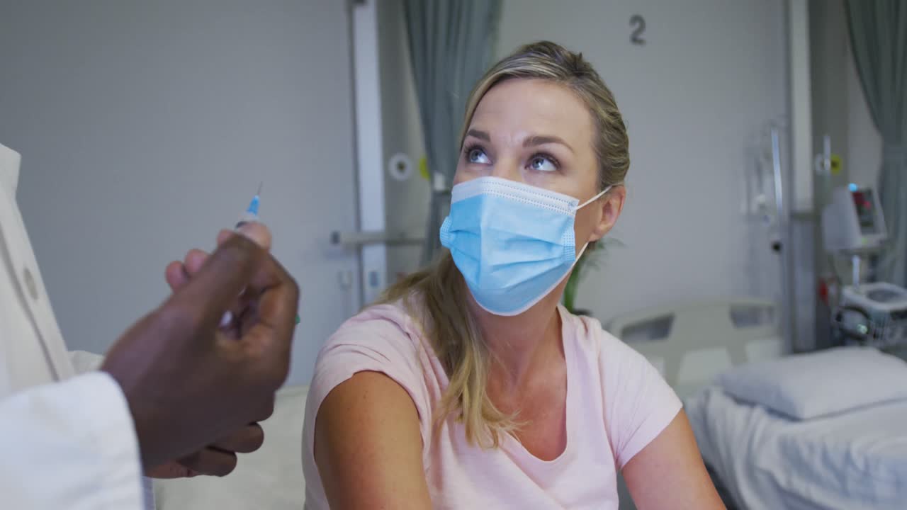 Eine kaukasische Patientin mit Gesichtsmaske sitzt im Krankenhausbett und wartet auf eine Injektion.