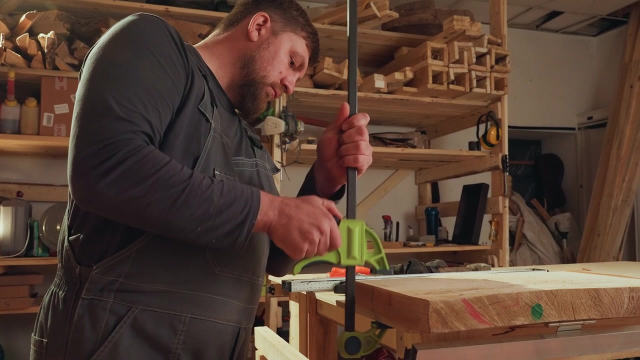 Man in overalls secures wood piece with green clamp on workbench surrounded by woodworking tools and materials including sawdust power tool measuring tape hand tools lumber clamps workshop