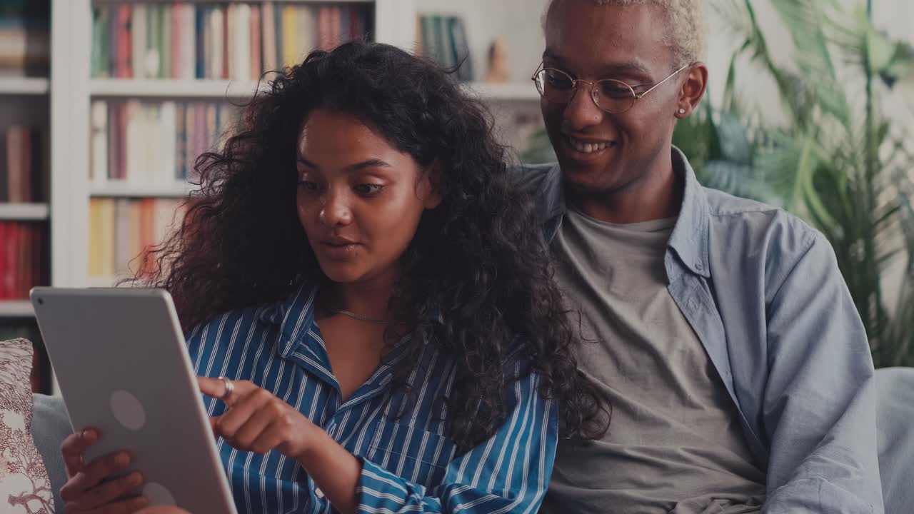 African boyfriend and indian girlfriend relaxing sit on couch holding tablet