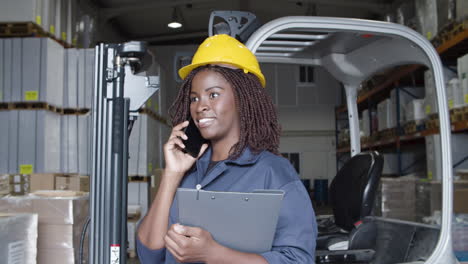 trabajadora afroamericana hablando por teléfono, de pie en el almacén y sosteniendo un portapapeles