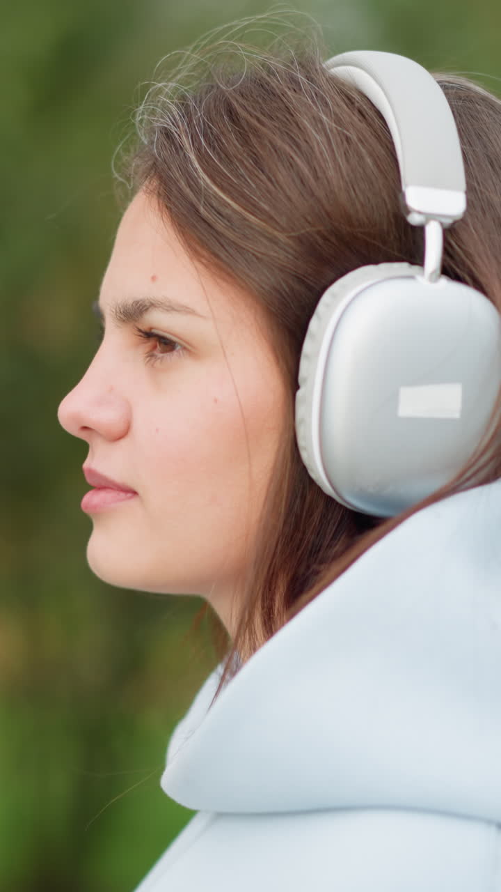 Close-up side view of young woman with headphones, listening to music outdoors in a garden with a blurred background of trees, perfect for videos on relaxation, music, or nature enjoyment