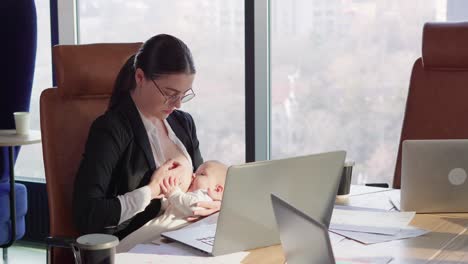 Una-Chica-Morena-Segura-De-Sí-Misma-Con-Gafas-Redondas-Y-Un-Uniforme-De-Negocios-Se-Sienta-En-Una-Mesa-Frente-A-Una-Computadora-Portátil-En-La-Oficina-Y-Amamanta-A-Su-Bebé-Sin-Distraerse-Del-Trabajo-En-Una-Oficina-Moderna.