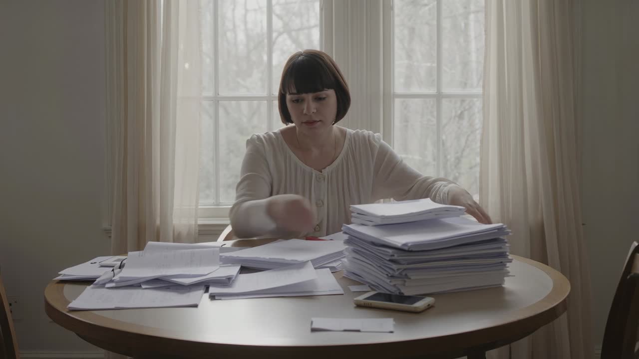 Seeing loose envelopes, woman wearing light top sorting papers at home table to tidy with phone