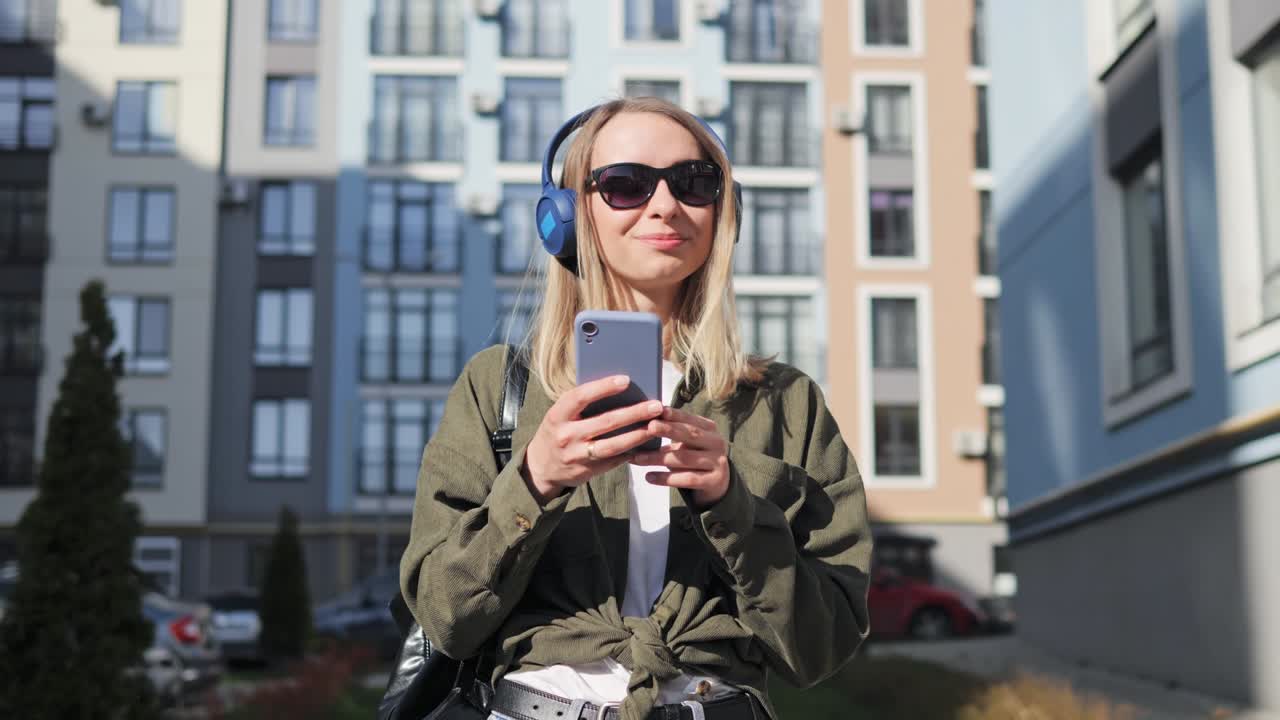 atractiva turista femenina caminando por el distrito de la ciudad escuchando música y escribiendo mensajes a amigos. teniendo tiempo libre en el fin de semana de verano. mujer navegando por internet. relajada. de buen humor. satisfecha.