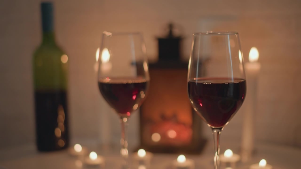 Duas taças de vinho tinto repousam sobre a mesa, com uma em foco nítido no primeiro plano e a segunda taça, garrafa e lanterna suavemente desfocadas ao fundo; a luz quente de velas cria reflexos dourados, um brilho suave e.