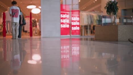 low-angle view inside mall showing people standing near bright red sale signs, with leg view of a lady in white canvas shoes passing by, reflections on shiny floor