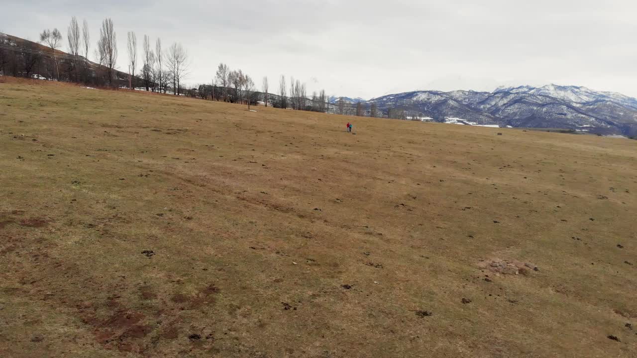 imágenes de drones de dos personas caminando a través de un campo abierto en un día soleado, con las montañas armenias en el fondo