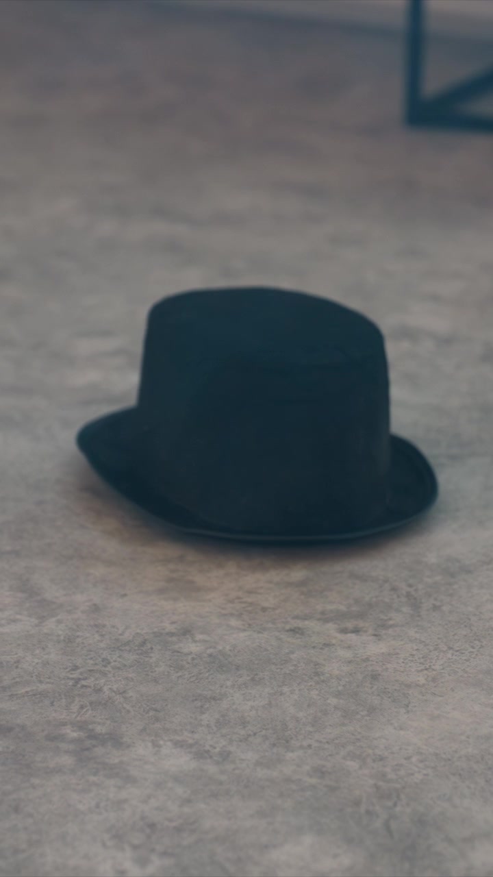 Black top hat spins and rolls on gray concrete floor. Dark metal structure is visible in upper corner of frame. Background includes partial view of industrial style frame