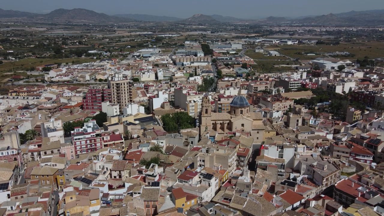 Filmagem aérea estática de drone mostrando o centro histórico da cidade de Aspe, Espanha, com foco na Basílica de Nuestra Señora del Socorro, com telhados e arquitetura clássicos espanhóis sob a luz do dia