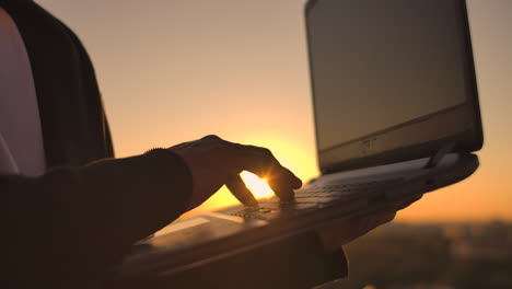 close-up: a programmer's hand typing on a laptop keyboard at sunset overlooking the roof. a businessman works remotely. freelancer performs work on vacation