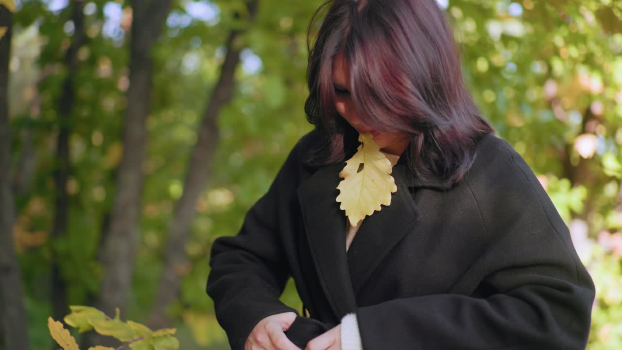 가을을 사랑하는 사람이 마른 잎을 턱 근처에 들고 코트 벨트를 조인 다음, 찻잔을 들기 위해 잎을 들어 올리는 모습이 확대되었어요. 부드러운 빛, 녹색 덮개, 조용한 길, 흩어진 나뭇잎이 있는 평온한 숲속 풍경이에요.