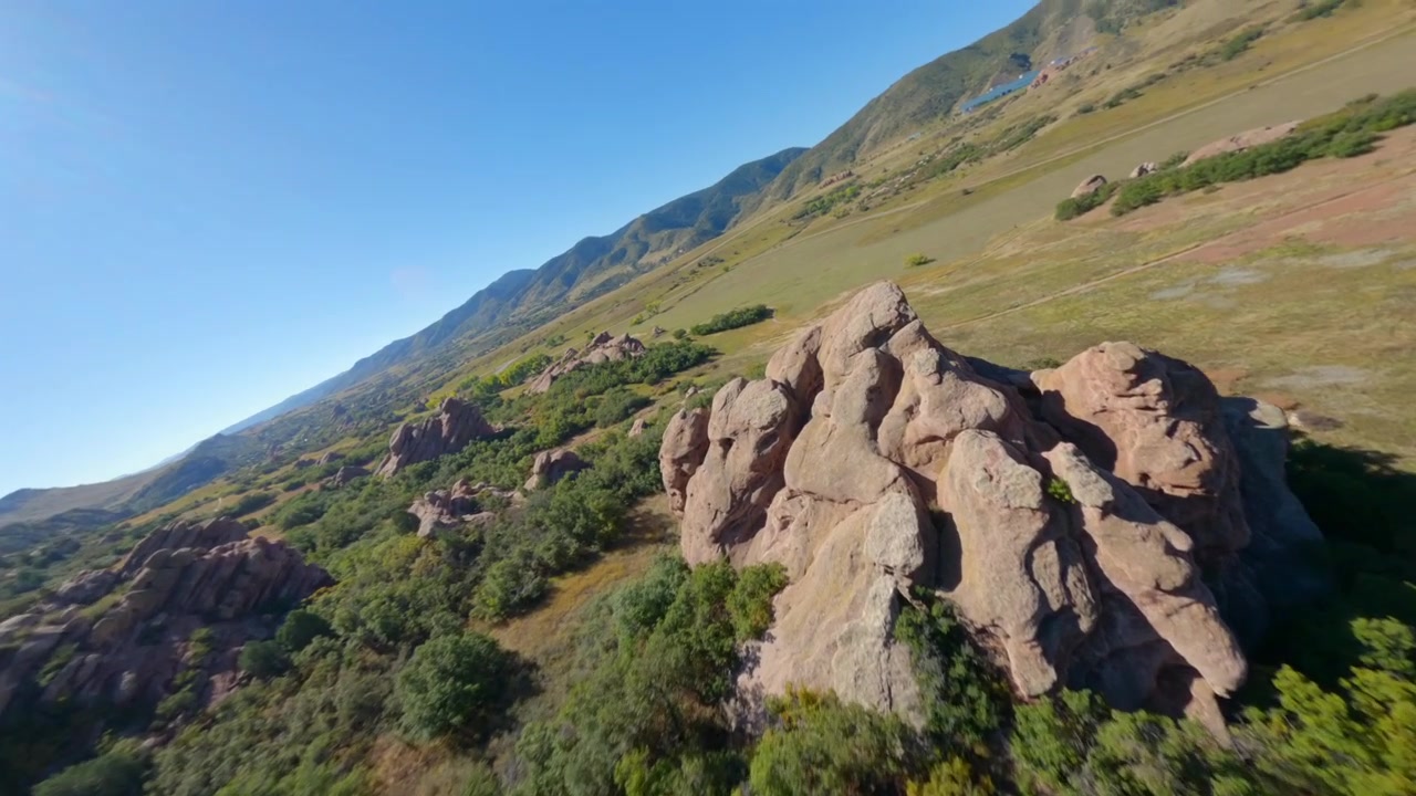 Affascinante filmato con drone FPV che vola basso sopra formazioni rocciose rosse uniche, simili a quelle aliene, immerse in un vibrante campo verde, mostrando paesaggi surreali e mozzafiato
