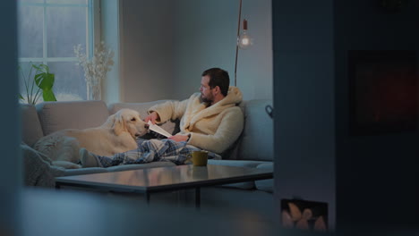 man with dog relaxing indoors, reading a book on a cold winter morning