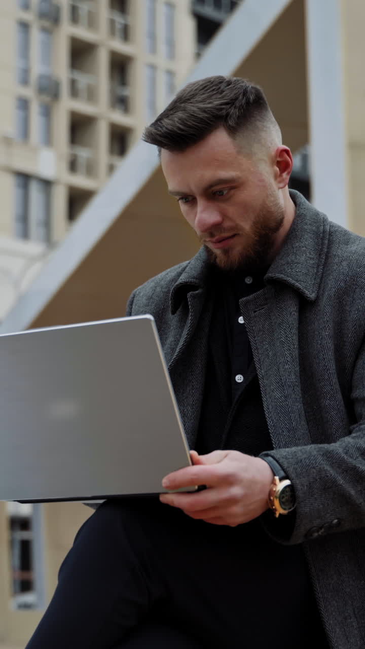 Young man with a laptop on fresh air. Handsome young man in a coat sitting in the street and working on a modern device in the city. Distance work. Vertical video