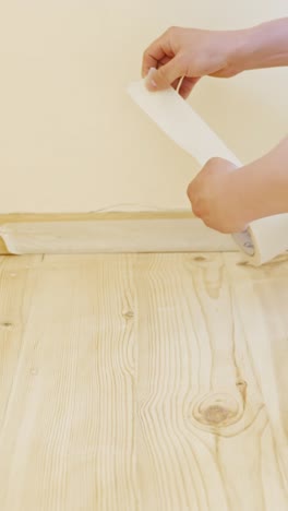 vertical close-up view showing hands applying paper tape along the baseboard of a light wooden floor, part of a house renovation process focused on clean edge preparation before painting