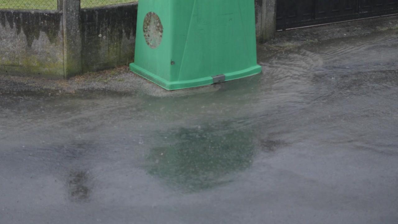 Close-up shot of torrential rain pouring on city pavement, water pooling and flooding around a green bin, capturing stormy, dramatic weather.