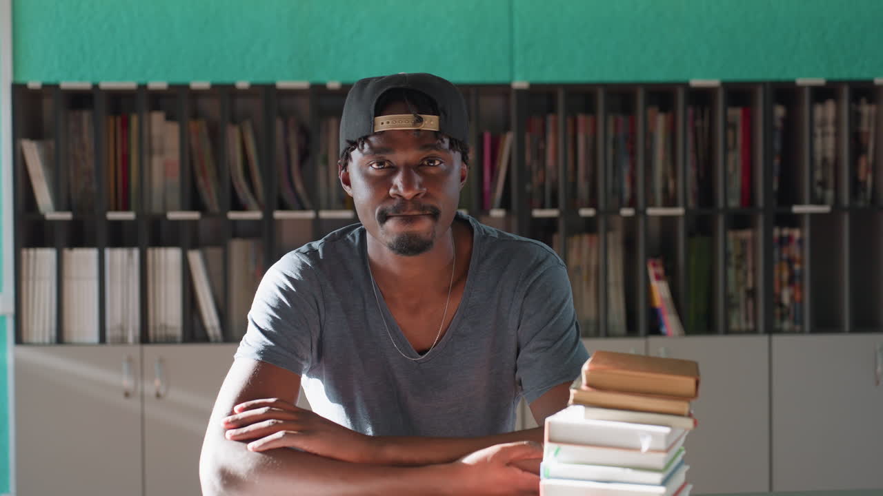 Young man wearing cap sits at library table with crossed arms, looking directly at camera next to stack of books, teal wall and shelves behind him, sunlight casting soft glow on serene academic scene