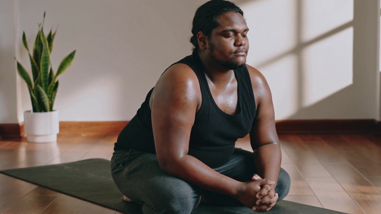 Settling man adjusting on mat for meditation in black tank and pants, with plant, window shadow
