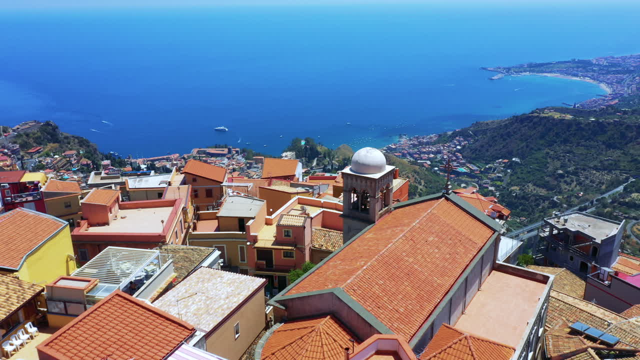 Aerial - drone flies over a small Sicilian village located on a cliff and reveals breathtaking view of green mountains with numerous houses and blue Mediterranean Sea in the background. Sicily, Italy.