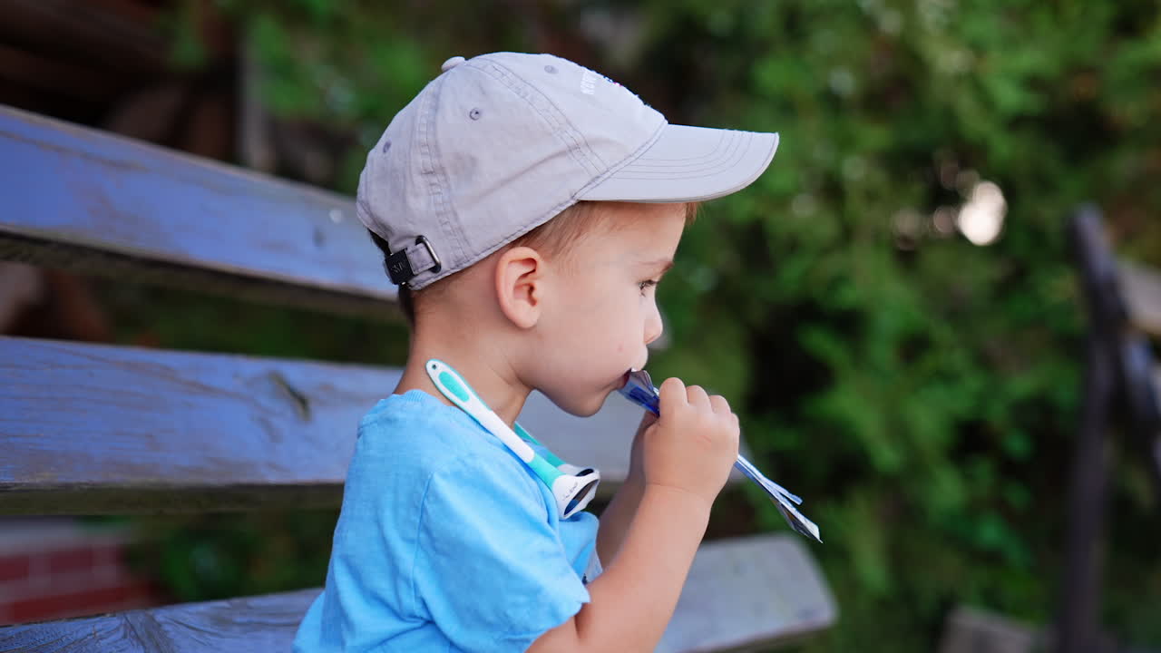 Caucasian toddler in blue t-shirt and grey cap sits on the bench outdoors. Baby drinks juice from pack and coughs. Side view close up.