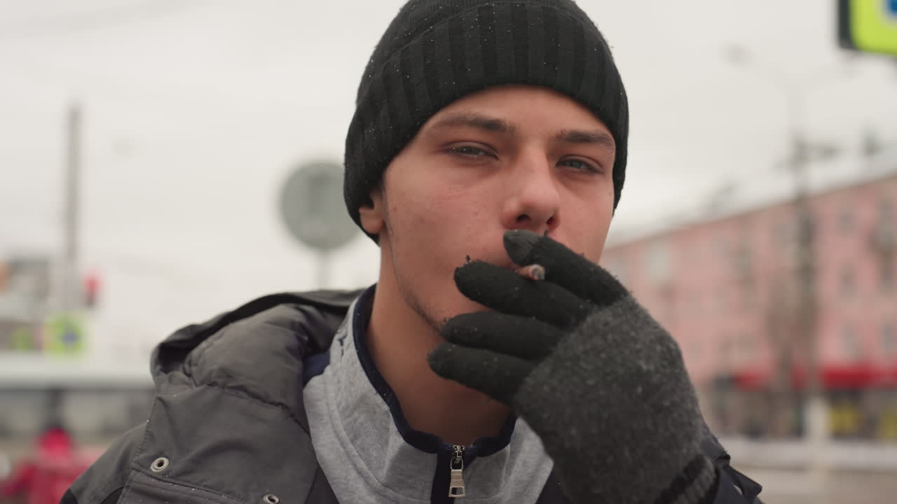 Ein nachdenklicher Jugendlicher macht draußen eine Pause mit Zigarette. Eine urbane Szene zeigt einen jungen Mann, der an einem kühlen Tag auf den Bus wartet. Der Teenager steht an einer Bushaltestelle unter bewölktem Himmel mit nachdenklichem Ausdruck.