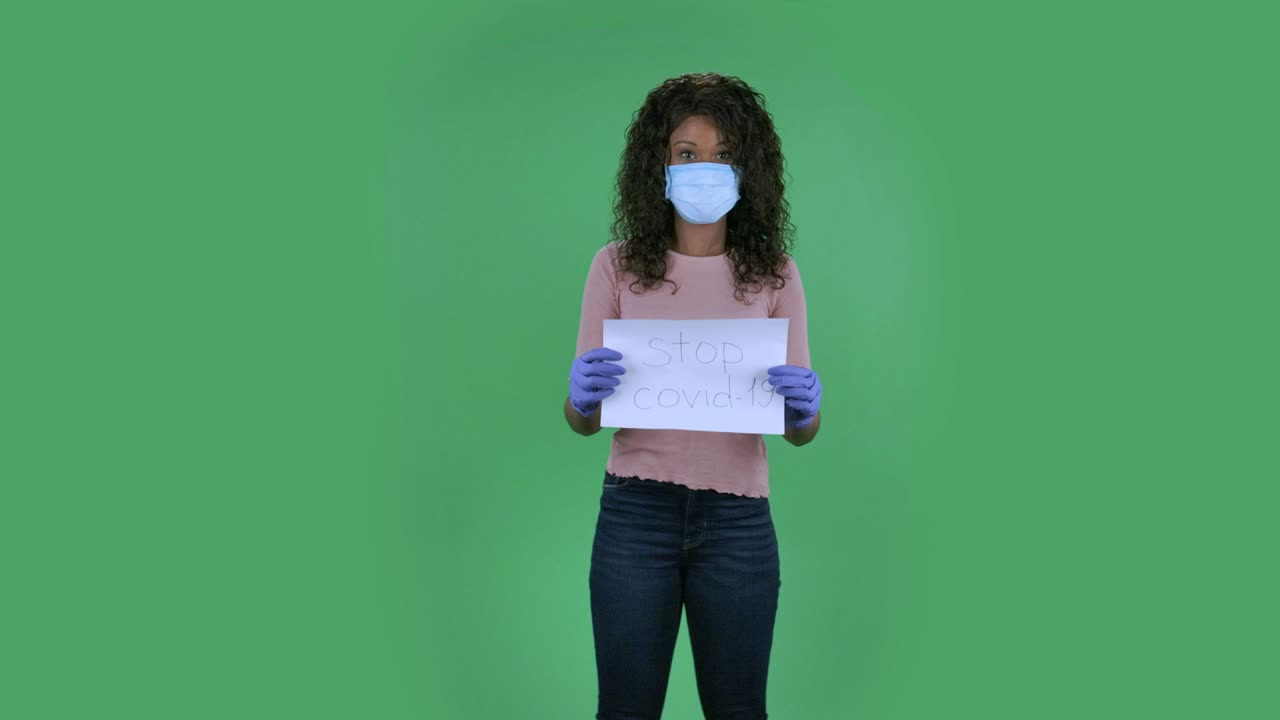 retrato de una hermosa joven afroamericana con máscara médica mirando a la cámara y sosteniendo un cartel de detención de covid-19, una morena ardiente con cabello ondulado en vaqueros y una blusa beige en una pantalla verde en el estudio.