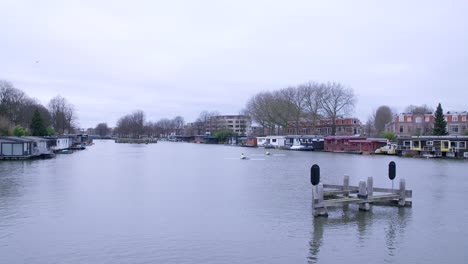 Wideangle-clip-of-two-boats-rowing-along-Utrecht's-canal-on-a-grey-cloudy-day,-with-traditional-waterside-houses-lining-the-banks-of-the-water