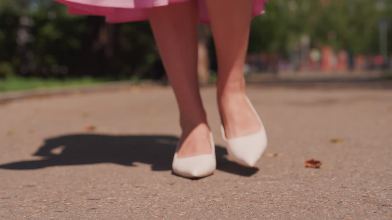 Scarpe bianche che camminano su un marciapiede in primo piano, ballerine a punta che picchiettano sul cemento, orlo della gonna che ondeggia, ombra marcata, passi ritmici lungo il sentiero soleggiato del parco, momento di moda casual e composto