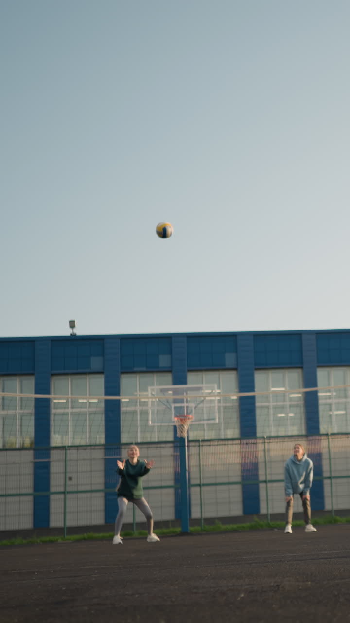 볼리볼 코트의 반대편에 두 명의 여성이 있습니다. 녹색 레을 입은 여자가 볼리볼을 제공하면서 녹색 후디를 입은 여인이 그것을 잡기 위해 점프합니다. 건물과 야외 코트는 배경에서 볼 수 있습니다.