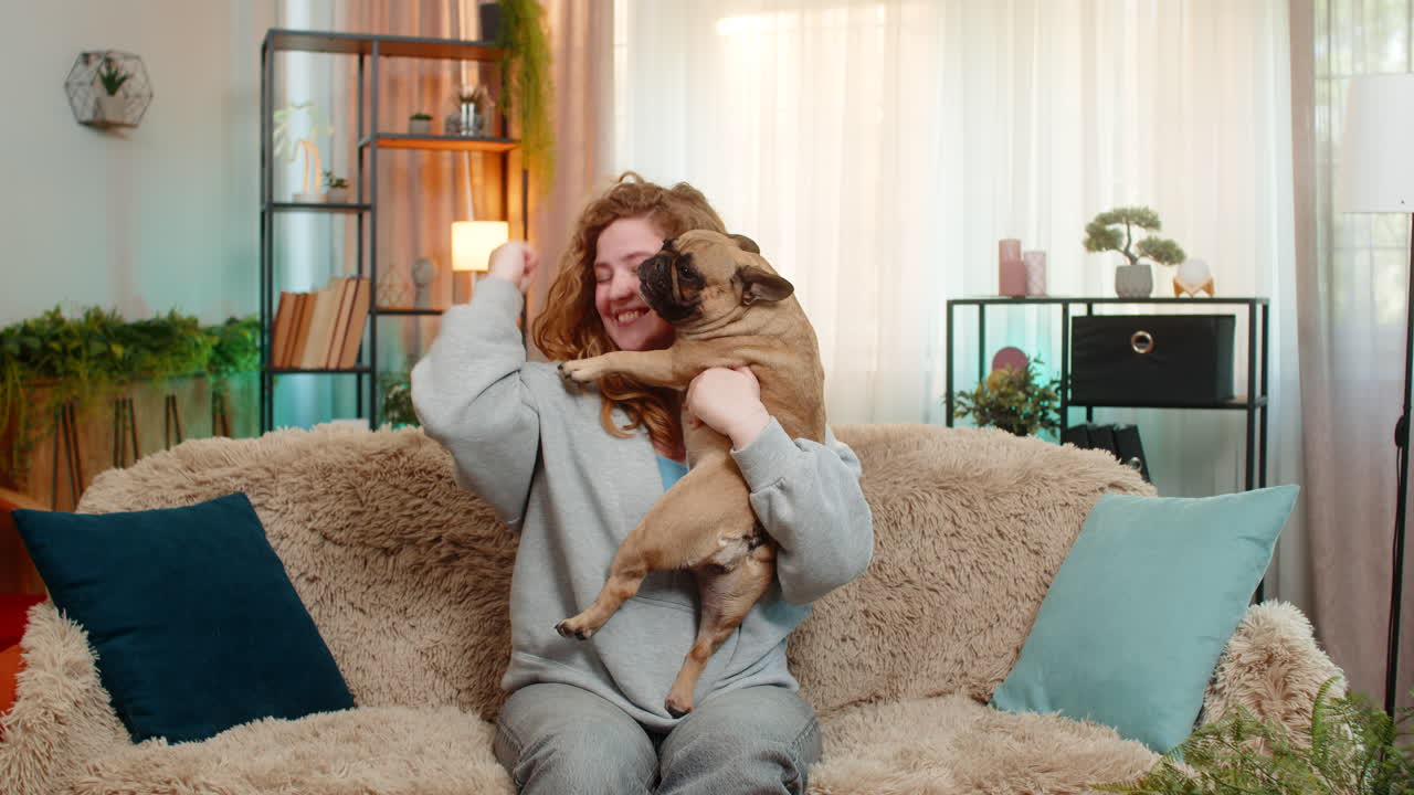 Young woman lifts pug dog with joy on sofa celebrating success and sharing happy cheerful emotion