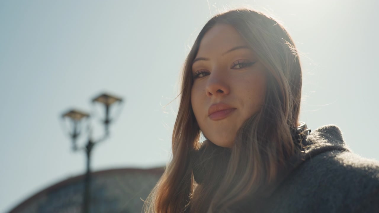 Donna elegante illuminata dal sole. Serena signora asiatica che sistema le cuffie in una luminosa piazza. Scena di strada pacifica con una donna alla moda che sintonizza le cuffie alla luce del sole con effetti silhouette.
