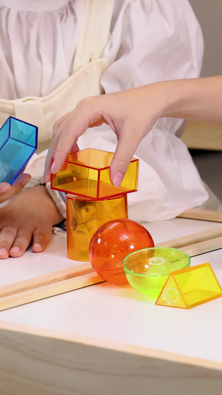 Mãos de criança e adulto empilham blocos geométricos coloridos em uma mesa de sala de aula sob iluminação brilhante.