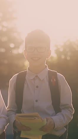 funny schoolboy in glasses throws pile of papers walking with friend after lessons in park at back sunlight slow motion