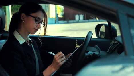 atractiva mujer de negocios caucásica en un coche estacionado, escribiendo notas en un clipboard, mientras se concentra y piensa
