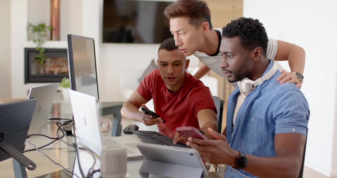 Diverse male coworkers walking in leaning over monitor and laptop sharing phones at glass desk