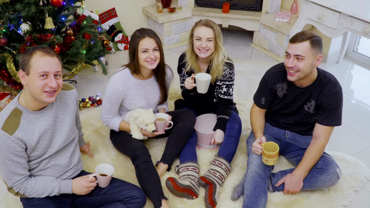 네 명의 젊은이들이 크리스마스 트리 근처의 바닥에 앉아 차나 커피를 마시고 있습니다.