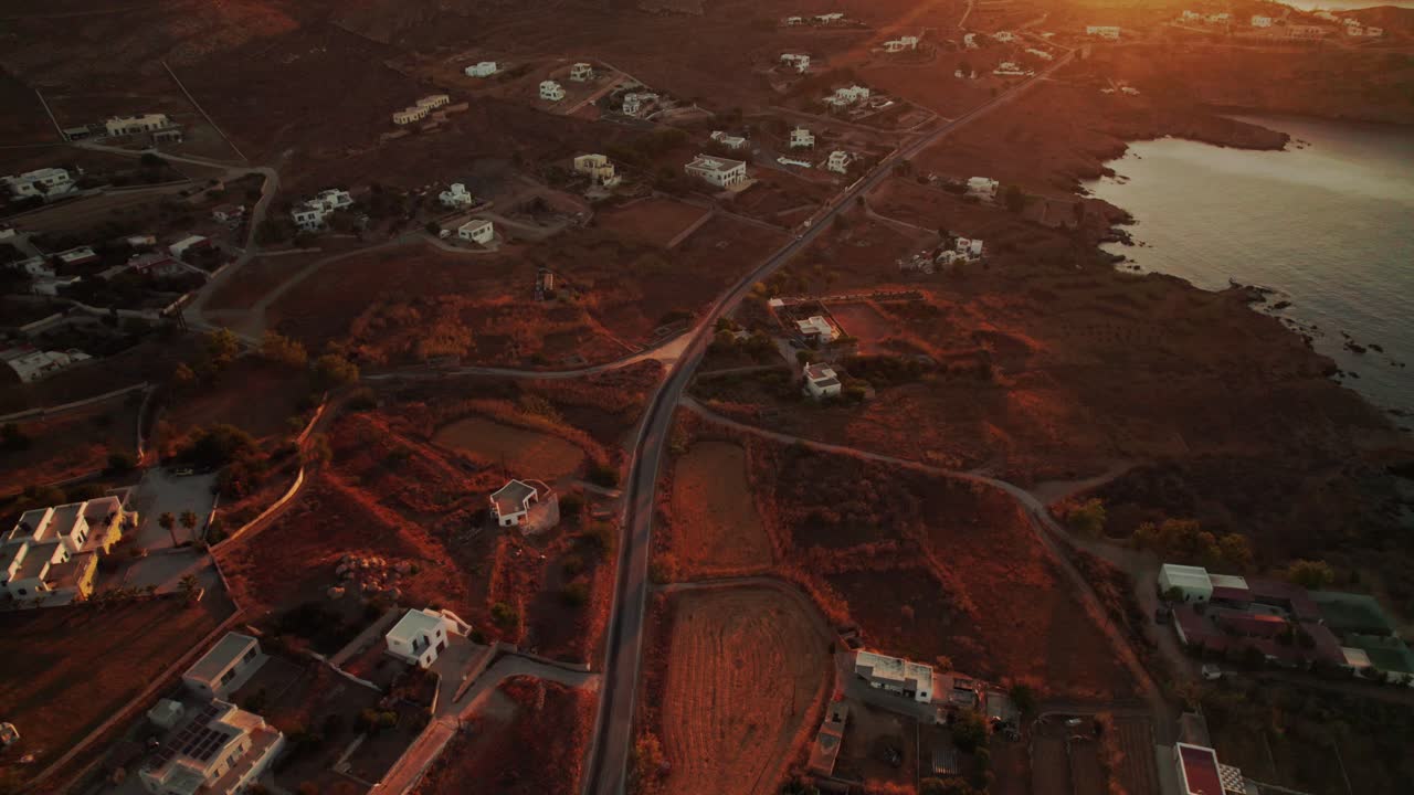 Drone bird's eye view above winding coastal road tilts up to yellow sunset, syros greece
