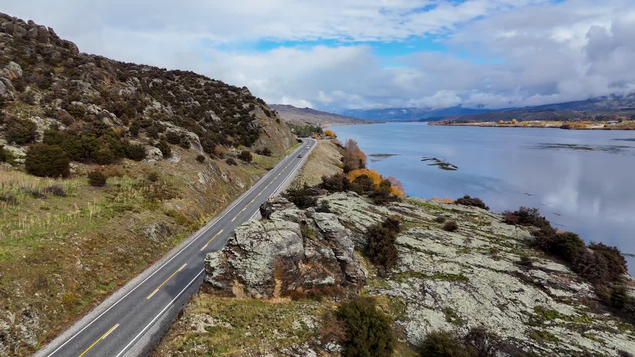 Drone camera glides above a winding lakeside road, passing rocky terrain and reflecting water under bright, natural daylight with sweeping cinematic movement