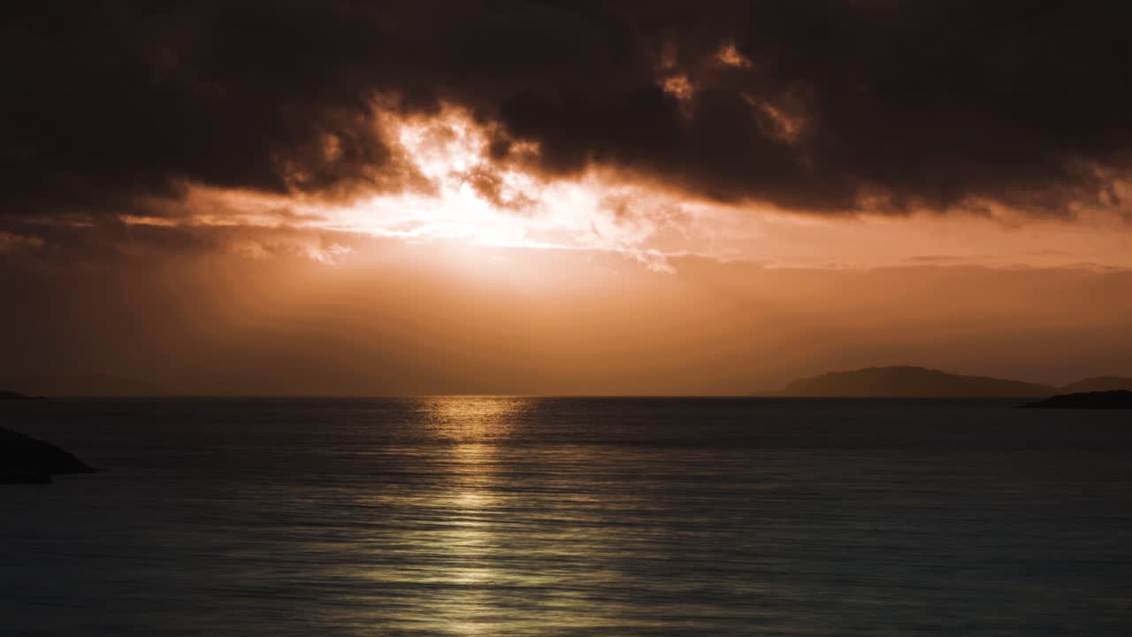 As the sun sets, the tranquil waters reflect the warm orange and red hues of the sky and distant islands.