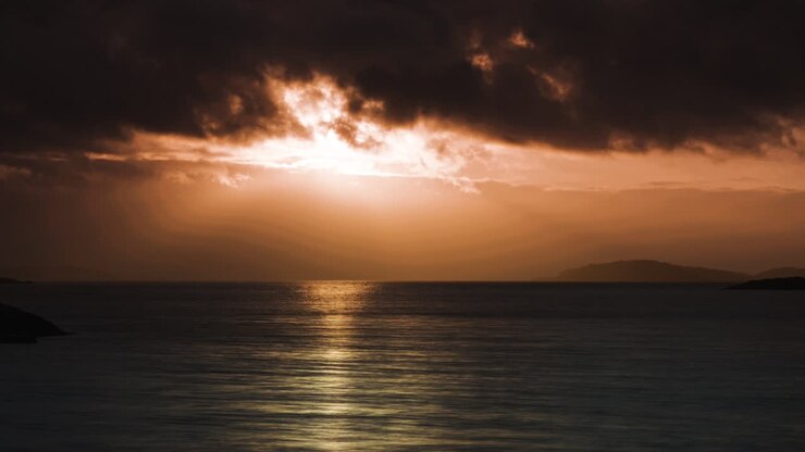 As the sun sets, the tranquil waters reflect the warm orange and red hues of the sky and distant islands.