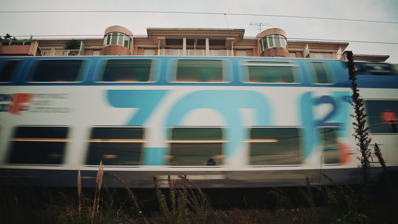 A blue commuter train speeds past a residential building, creating strong motion blur against the still architecture