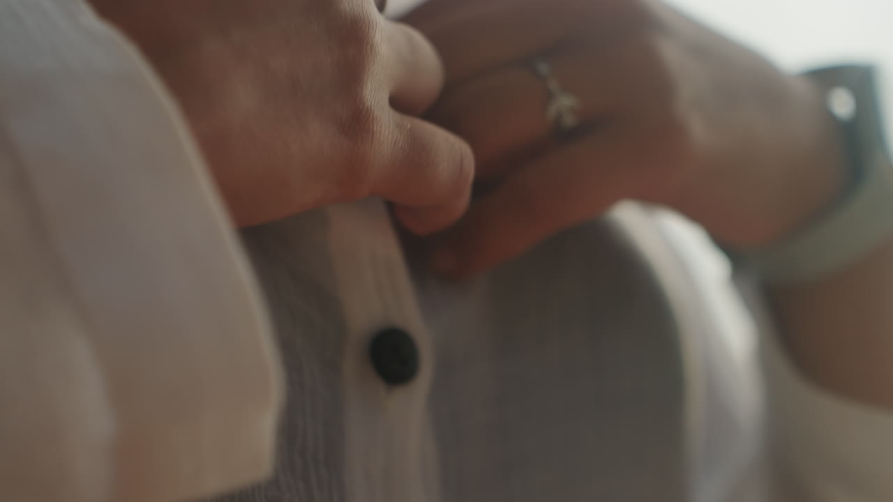 Tangan mengancingkan blus tipis dari dekat, persiapan pagi yang percaya diri dengan wanita menyesuaikan pakaian sebelum keluar, sedikit memperlihatkan garis leher di bawah linen, cahaya latar hangat menyoroti gerakan jari yang mengamankan.