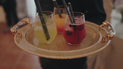 three colorful cocktails served in glasses with straws, presented on a gold tray with elegant handles